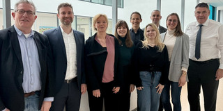 9 Personen verschiedenen Alters und Geschlechts in Smart Causal gekleidet stehen lächelnd nebeneinander auf einem Büroflur.