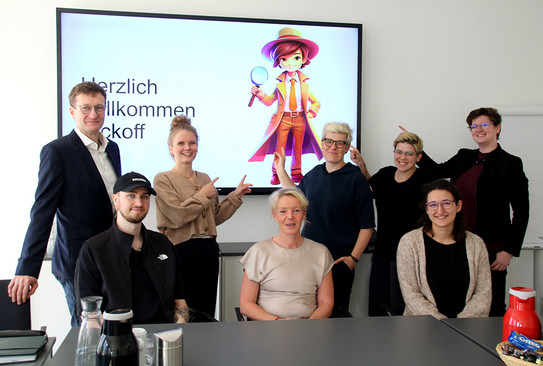 Acht Personen unterschiedlichen Alters und Geschlechts stehen vor einem Präsentationsbildschirm. Mehrere Personen zeigen auf die gezeigte Präsentationsfolie. Die Folie zeigt einen animierten Detektiv mit Lupe.