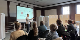 Personen sitzen in einem Konferenzraum. Ein Referent steht vor einer Leinwand-Präsentation.